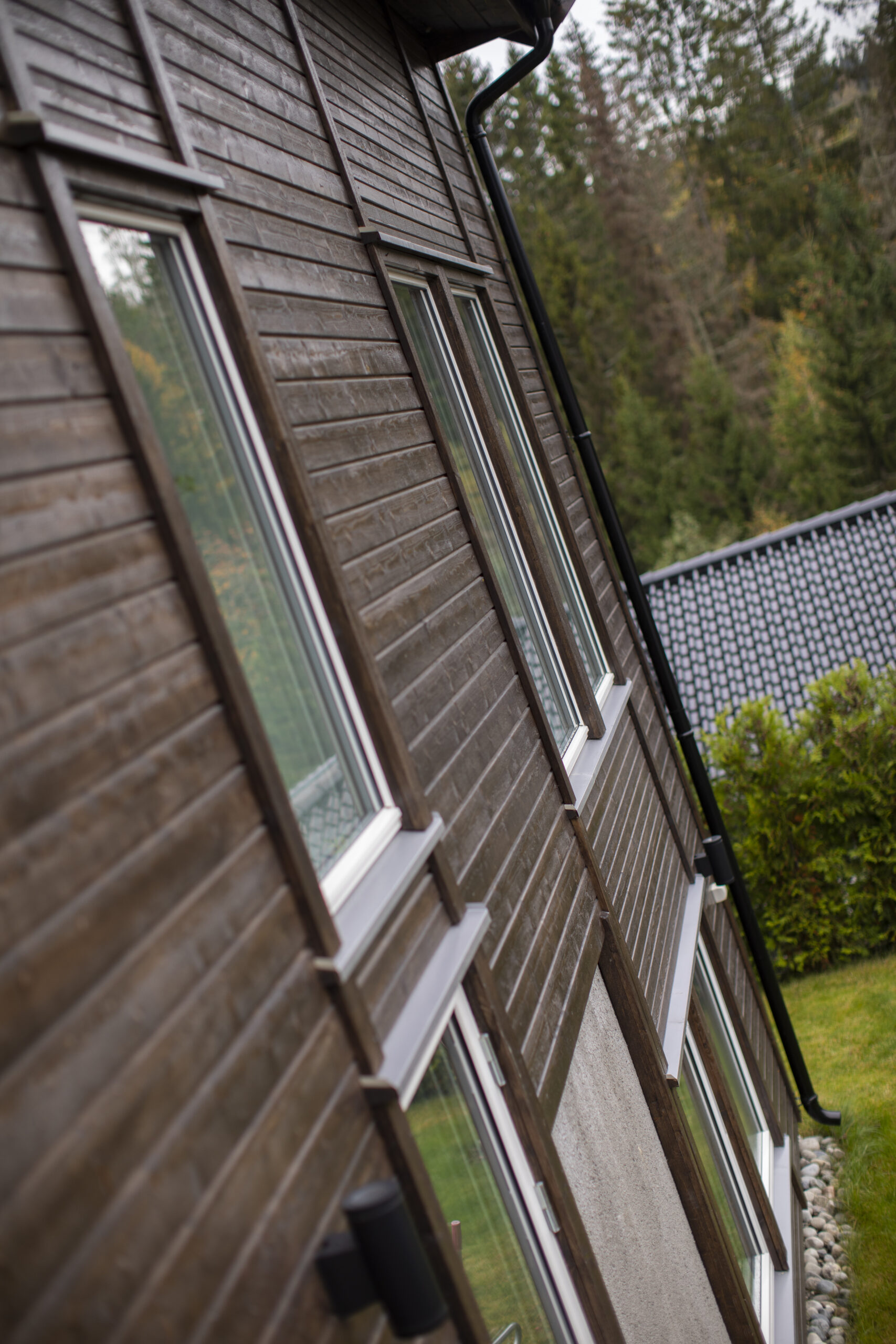 Brown wooden house exterior with white-framed windows and a black gutter, photographed at an angled perspective.